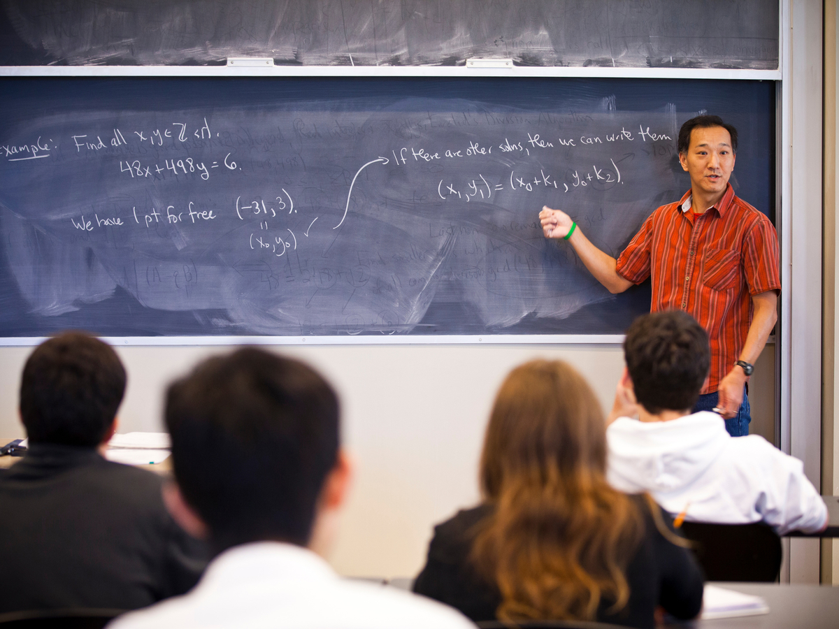 Asian faculty member standing in front of a chalkboard with formulas written in chalk. Students are blurred out in the foreground of the photo facing the teacher, so you only see the backs of their heads.