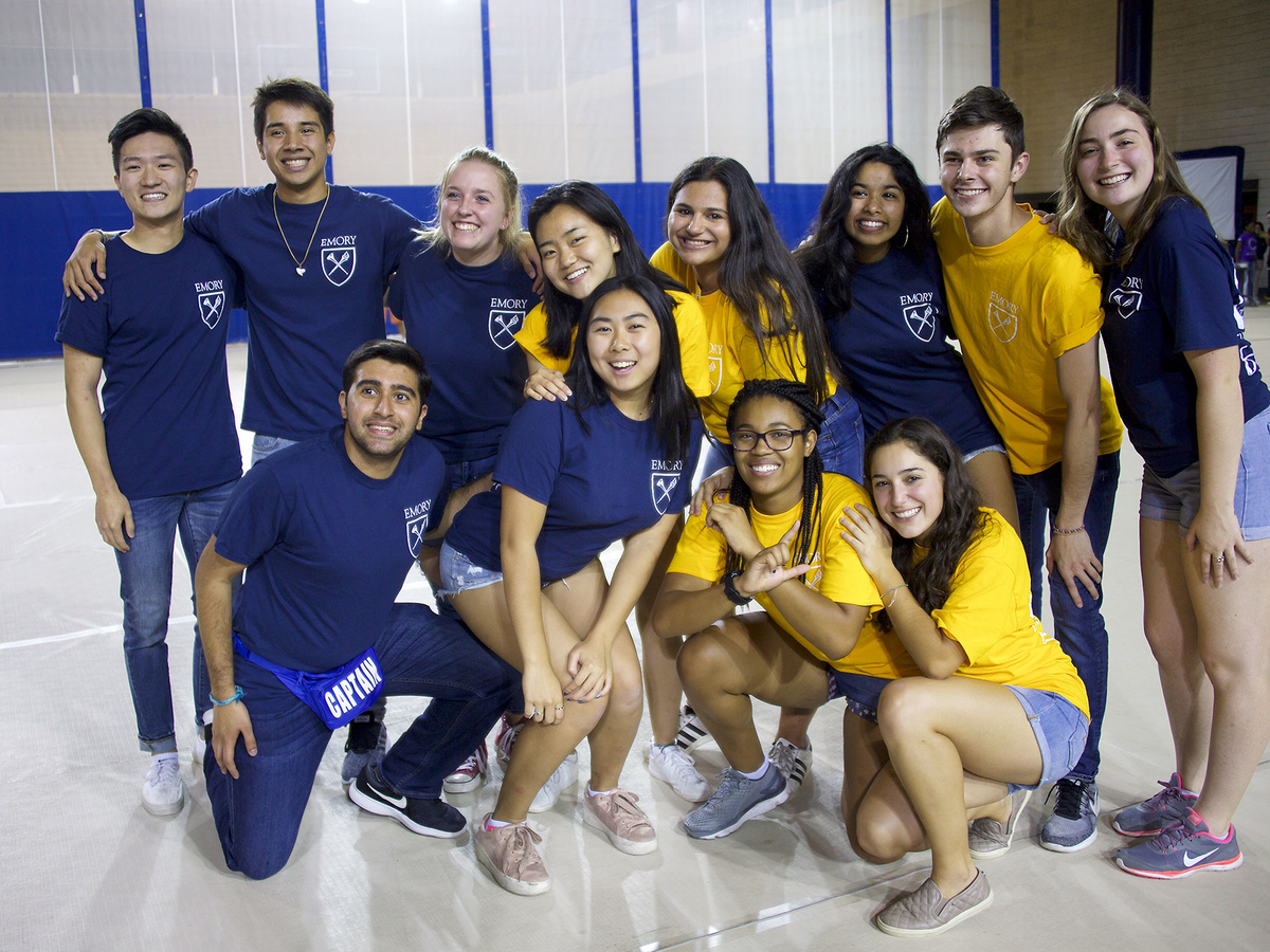 A group of 12 undergraduate students standing together in a gym looking at the camera. Half of the students are wearing blue Emory t-shirts while the other half are wearing gold Emory t-shirts.