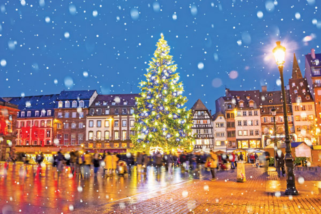An adorned Christmas tree standing tall in the square of a European city. The square is lined with row-style houses on all sides. Snow is falling, which creates a distinctive holiday feel to the photo.