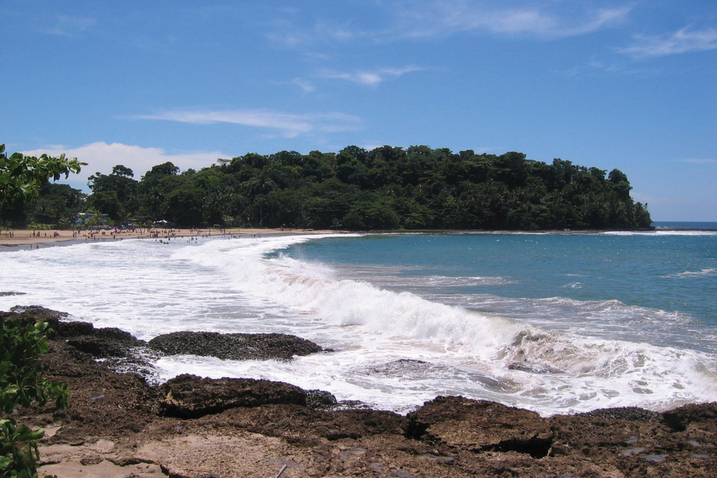 Photograph of a Costa Rican beach with waves crashing against a rocky shore with a dense treeline in the background.