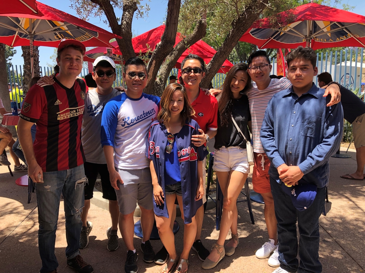 A co-ed group of alumni standing together outside in an patio with red umbrellas behind them. They're all looking at the camera and some of them are wearing sports fandom attire.