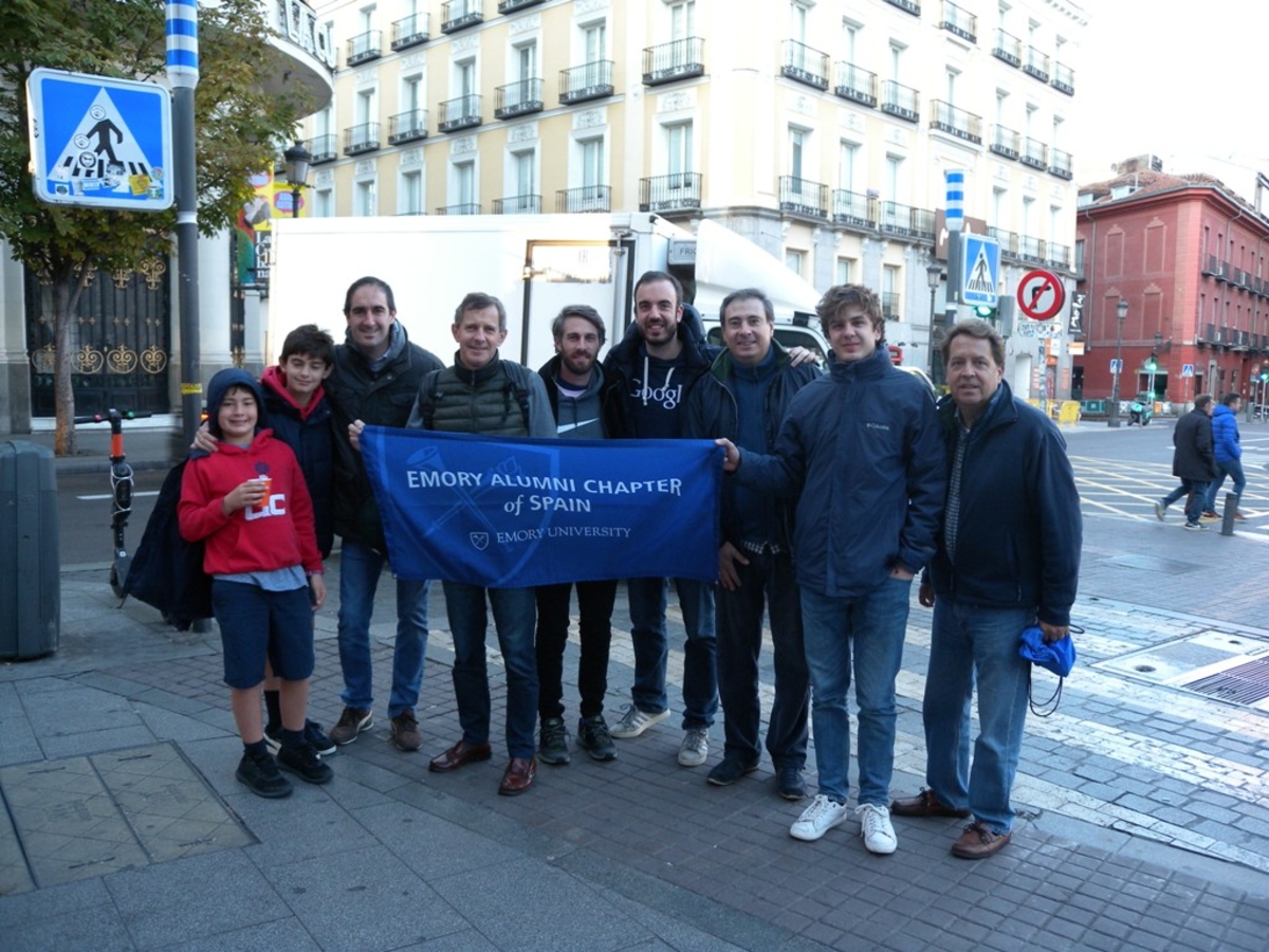 A group of seven male alumni with two young boys standing in a street. Each of them have coats, jeans, and tennis shoes on and are holding a flat that says Emory Alumni Chapter of Spain.