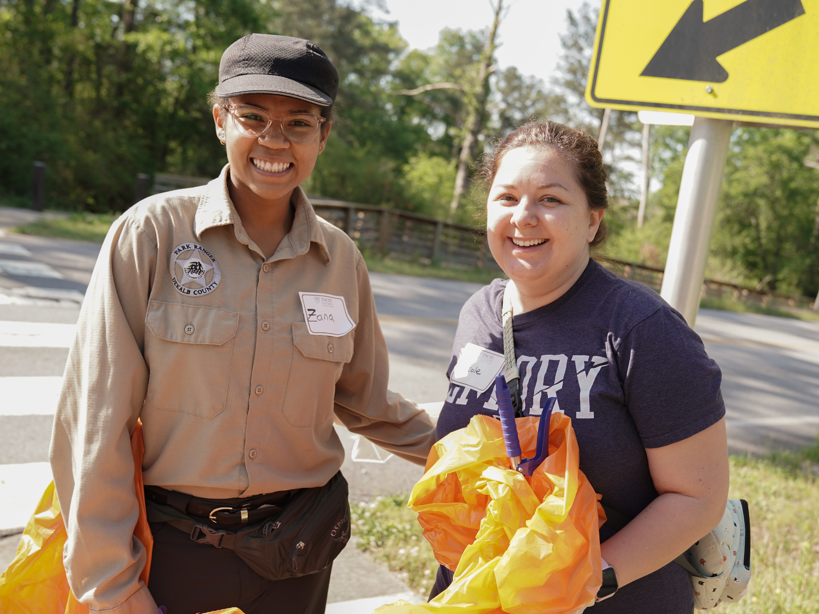 Two women standing together on the side of a road smiling at the camera while holding trashbags.
